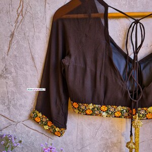 May include: A brown sheer blouse with a tie-up back and embroidered floral trim. The blouse has long sleeves and a V-neckline.