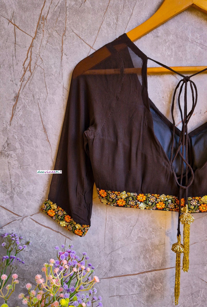 May include: A brown sheer blouse with a tie-up back and embroidered floral trim. The blouse has long sleeves and a V-neckline.