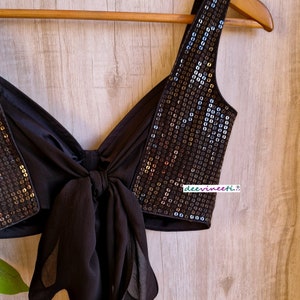 May include: A black sequined crop top with a bow tie detail. The top is on a wooden hanger and has a black fabric strap.