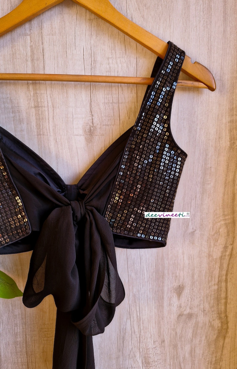 May include: A black sequined crop top with a bow tie detail. The top is on a wooden hanger and has a black fabric strap.