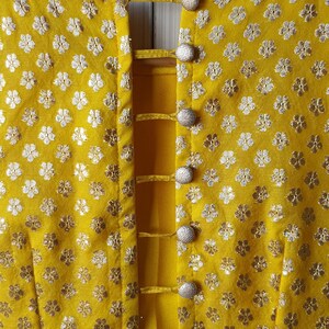 Pu&ograve; includere: Una blusa gialla con un dettaglio sul retro di una serie di cinturini e bottoni. La blusa ha un motivo floreale con fiori argentati su uno sfondo giallo. Il testo "deevineeti..." &egrave; visibile nell'angolo in basso a destra dell'immagine.
