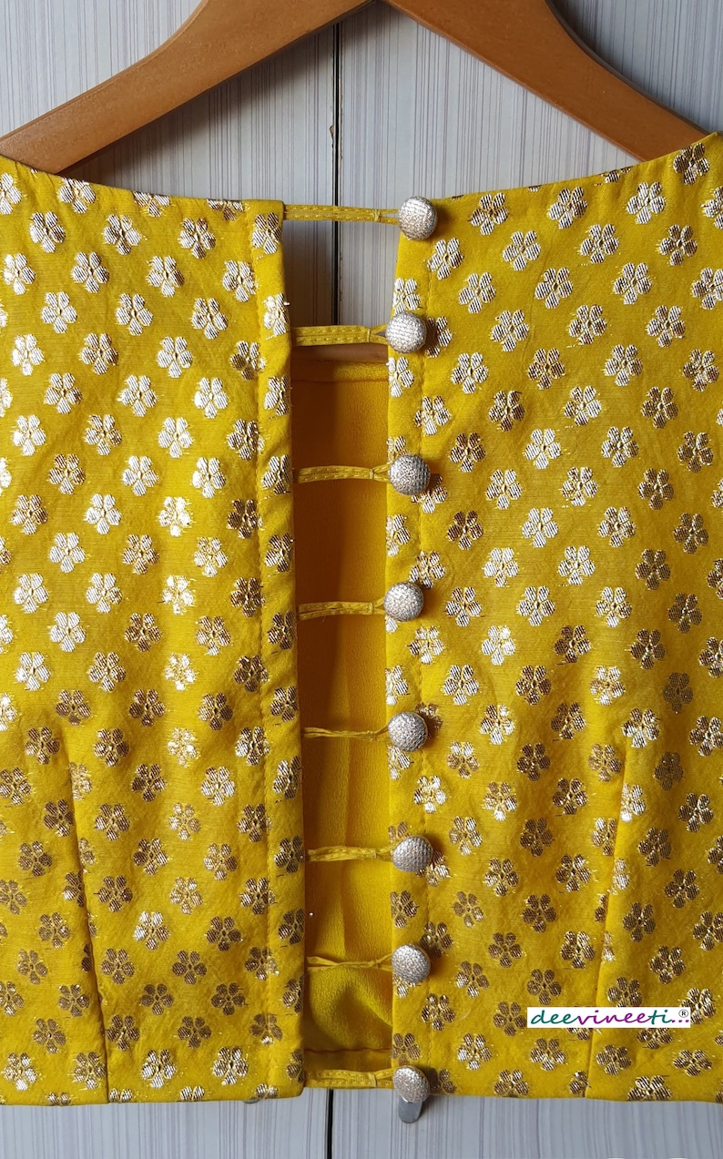 Pu&ograve; includere: Una blusa gialla con un dettaglio sul retro di una serie di cinturini e bottoni. La blusa ha un motivo floreale con fiori argentati su uno sfondo giallo. Il testo "deevineeti..." &egrave; visibile nell'angolo in basso a destra dell'immagine.