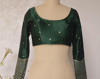 May include: A dark green long-sleeved blouse with a scoop neckline. The blouse has small gold embroidered details and gold trim on the neckline and hem. The sleeves feature a detailed gold pattern near the cuffs. The blouse is displayed on a mannequin.