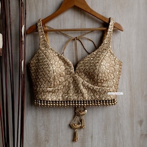 May include: Gold sequined top with a deep V-neck and a beaded hemline. The top is hanging on a wooden hanger. A decorative tassel hangs from the bottom of the top. The top is a formal wear item.
