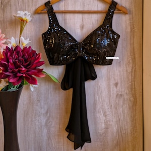 May include: A black sequined crop top with a bow tie detail in the front. The top has a deep V-neckline and thin straps. The bow tie is made of black chiffon.