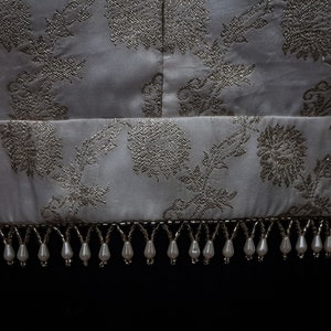 May include: A white top with a silver floral pattern and a beaded trim. The trim is made of white beads with silver accents.
