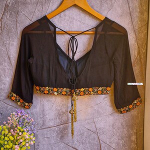 May include: Black sheer blouse with a tie-up back and floral embroidery around the neckline and sleeves. The blouse has a deep V-neckline and is made of a lightweight fabric.