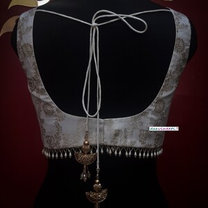 May include: White embroidered blouse with a white string tie and gold and pearl embellishments. The blouse has a deep V-neck and a cropped length. The text "deervineeti." is visible on the bottom right corner of the blouse.