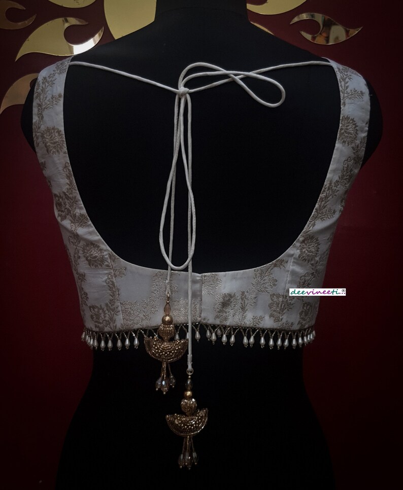 May include: White embroidered blouse with a white string tie and gold and pearl embellishments. The blouse has a deep V-neck and a cropped length. The text "deervineeti." is visible on the bottom right corner of the blouse.