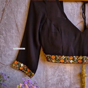 May include: Black sheer blouse with embroidered floral trim and a gold tassel. The blouse has a V-neckline and three-quarter sleeves. The text "deevineeti." is visible on the blouse.