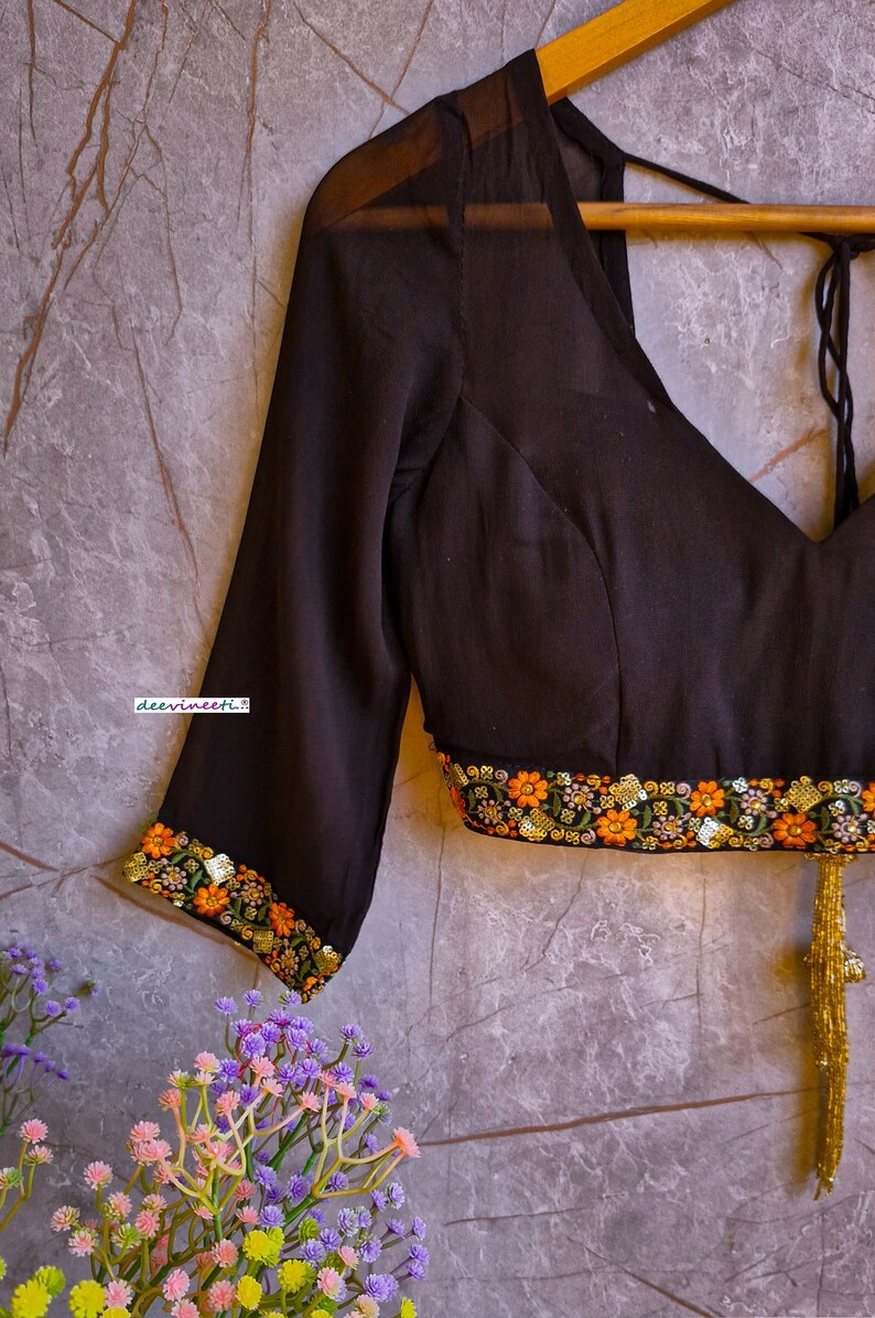 May include: Black sheer blouse with embroidered floral trim and a gold tassel. The blouse has a V-neckline and three-quarter sleeves. The text "deevineeti." is visible on the blouse.