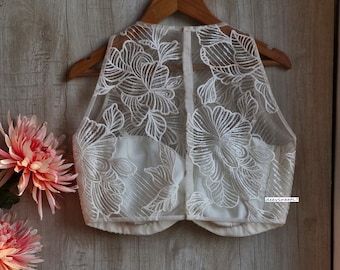 Puede incluir: Blusa blanca sin mangas con bordado floral. La blusa es transparente y tiene un diseño delicado y femenino.