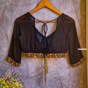 May include: A black sheer blouse with a deep V-neckline and a tie at the back. The blouse has a gold embroidered trim around the neckline and sleeves. The blouse is on a hanger against a textured gray background.