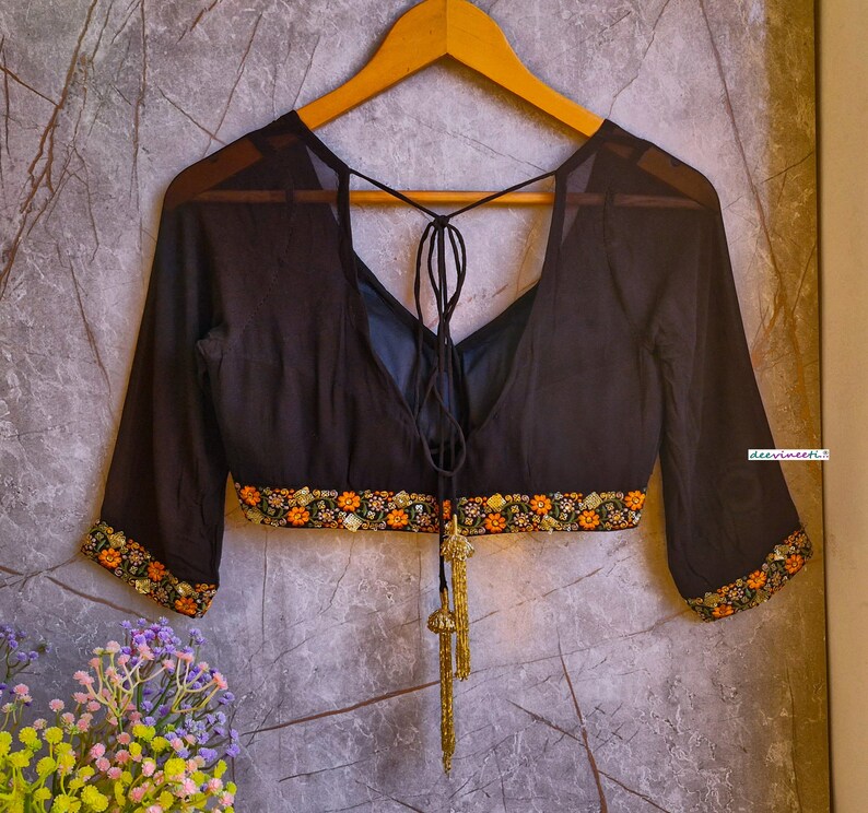 May include: A black sheer blouse with a deep V-neckline and a tie at the back. The blouse has a gold embroidered trim around the neckline and sleeves. The blouse is on a hanger against a textured gray background.