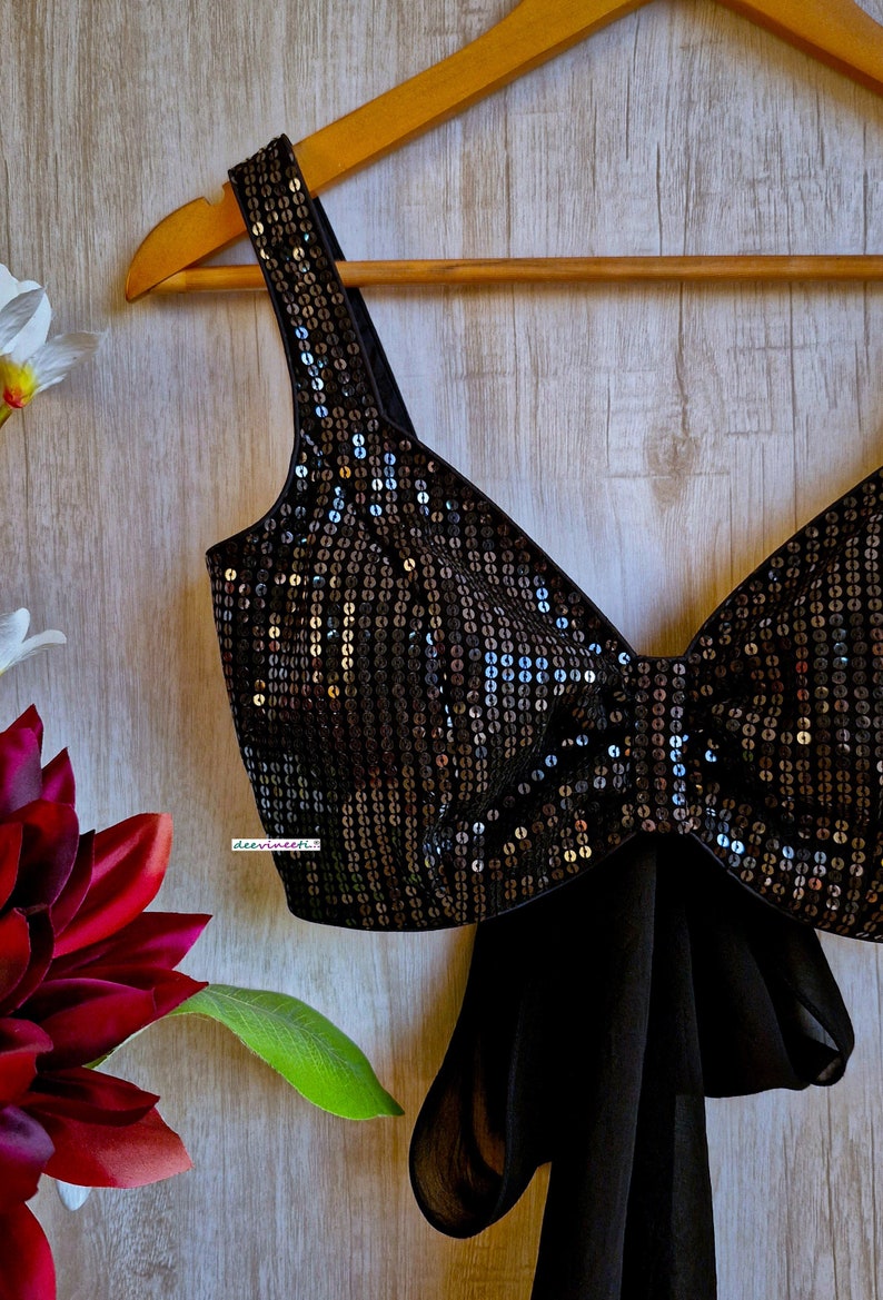 May include: A black sequined crop top with a bow detail and a matching black chiffon scarf. The top is on a wooden hanger.