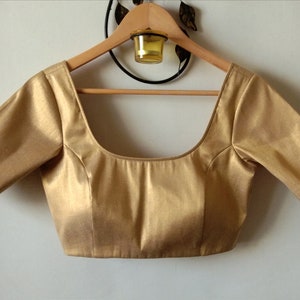 Puede incluir: Una blusa dorada de manga corta con escote redondo. La blusa está colgada de una percha de madera.