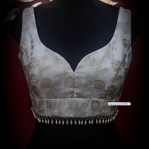 May include: A white sleeveless blouse with a silver floral pattern and a silver beaded trim. The blouse is on a black mannequin.