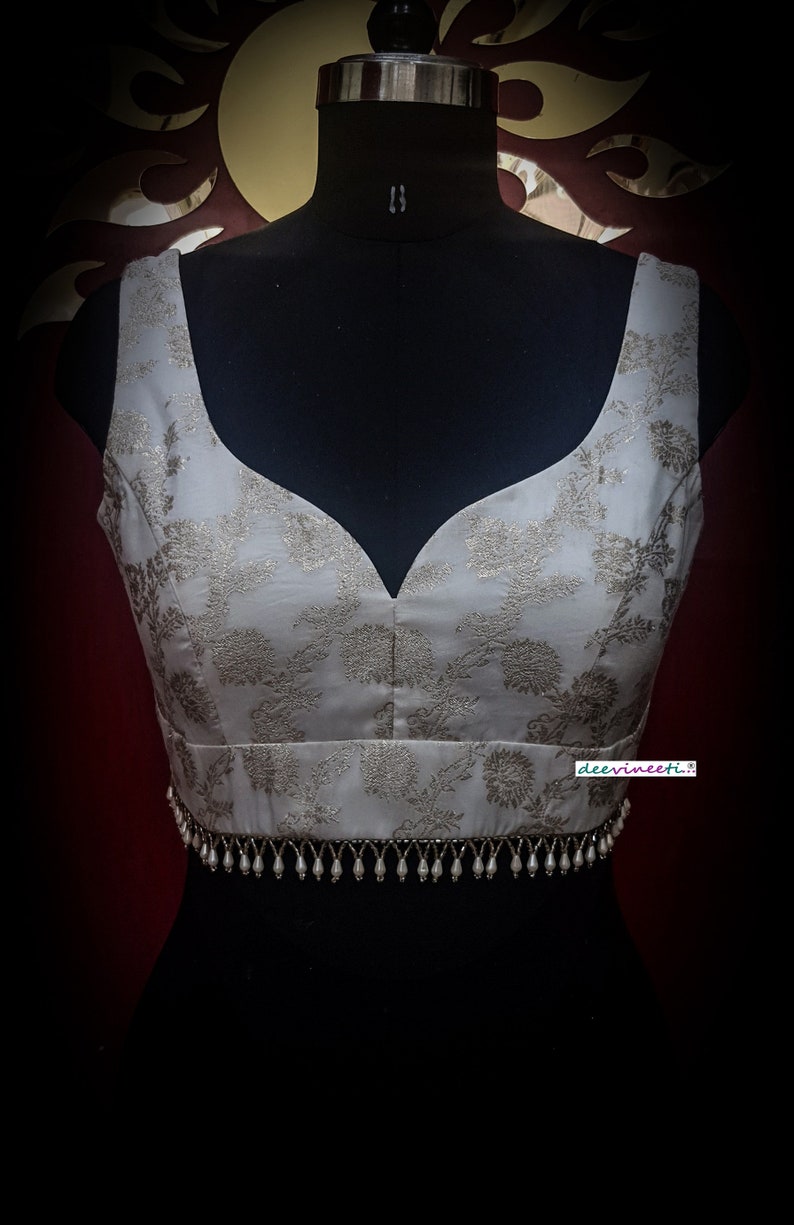 May include: A white sleeveless blouse with a silver floral pattern and a silver beaded trim. The blouse is on a black mannequin.