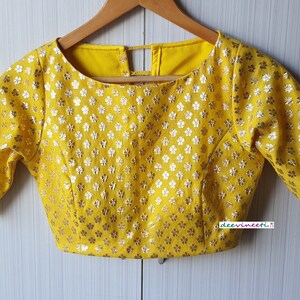Pu&ograve; includere: Una blusa gialla con un motivo floreale argentato e una scollatura rotonda. La blusa ha maniche corte e un'apertura sul retro.