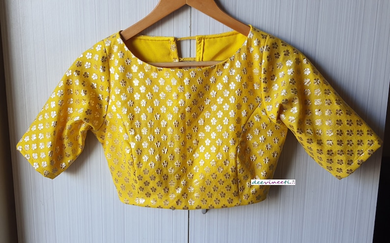Pu&ograve; includere: Una blusa gialla con un motivo floreale argentato e una scollatura rotonda. La blusa ha maniche corte e un'apertura sul retro.