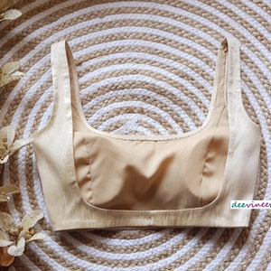 May include: A beige, sleeveless, padded bra with a scoop neck. The bra is made of a silky fabric and has a satin-like finish.