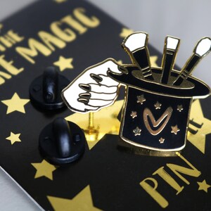 May include: Gold-tone enamel pin featuring a black top hat with three paintbrushes sticking out of it. A white hand with long nails is reaching into the hat. The hat has a gold heart with a gold outline and gold stars on it. The pin is on a black background with gold stars and the text "MAKE THE MAGIC" in gold letters. The text "PIN" is also in gold letters on the background.