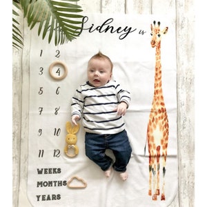 May include: A white baby blanket with a giraffe illustration, a baby lying on the blanket, and a number chart for tracking growth. The text "Sidney is" is written on the blanket.