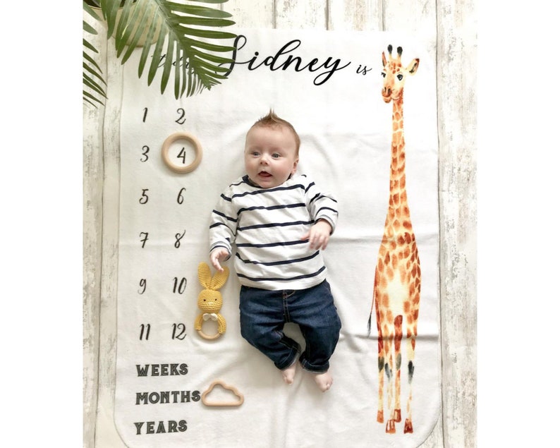 May include: A white baby blanket with a giraffe illustration, a baby lying on the blanket, and a number chart for tracking growth. The text "Sidney is" is written on the blanket.