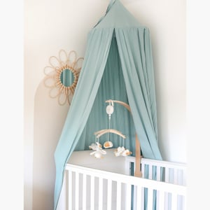 May include: A light blue canopy hangs over a white crib with a wooden mobile hanging from the top. The mobile has a wooden base and features white stuffed animals and a small wooden clock.