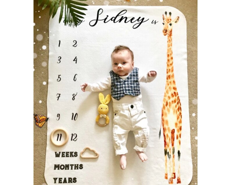 May include: A white baby blanket with a giraffe print and a baby lying on it. The blanket has numbers 1-12 printed on it, along with the words "Weeks", "Months", and "Years". The text "Sidney is" is printed at the top of the blanket.