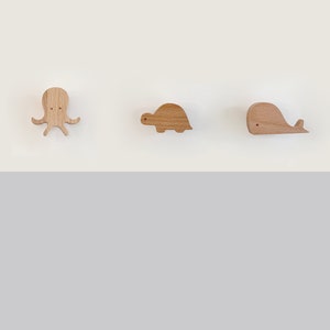 May include: Three wooden wall hooks shaped like an octopus, a turtle, and a whale. The hooks are a light brown colour and are mounted on a white wall.