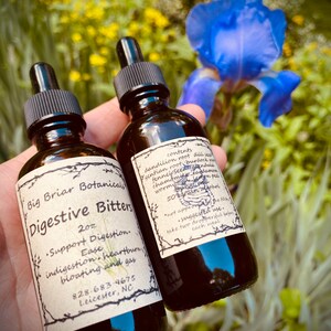 May include: Two brown glass bottles of "Digestive Bitters" with black droppers. The labels list ingredients and instructions. The bottles are held in front of a blurred background of flowers and greenery. The text on the label includes "Support Digestion" and "Ease indigestion, heartburn, bloating and gas."