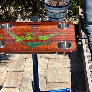May include: A wooden side table with a glossy finish, featuring four stainless steel cup holders. The table is decorated with a green and blue design. It has a blue metal base and is designed for outdoor use.