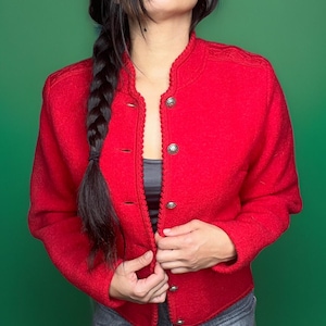 Puede incluir: Un cárdigan de lana rojo con ribete trenzado y botones plateados. El cárdigan está desabrochado y la parte delantera está abierta.