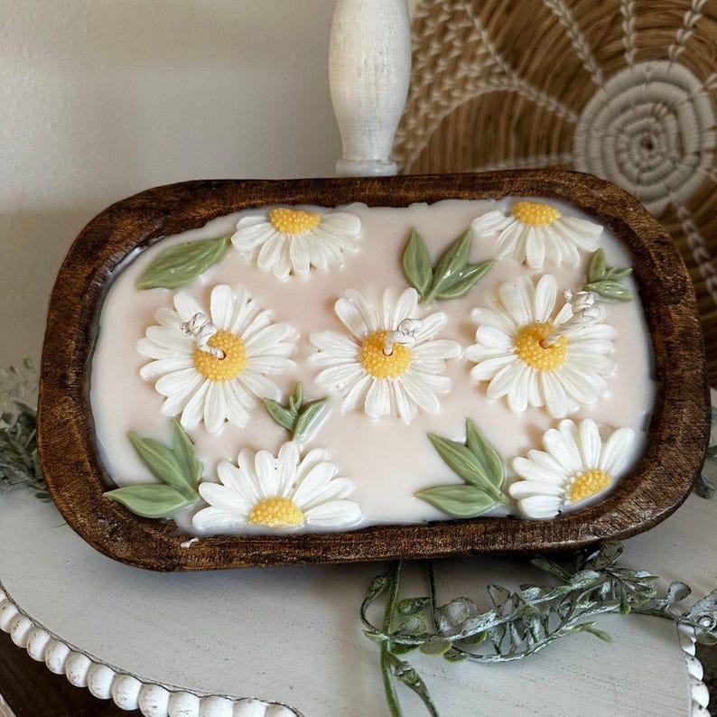 May include: A decorative candle in a wooden bowl. The candle features white daisy flowers with yellow centers and green leaves. The candle is set on a white surface.