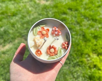 May include: A white candle in a round tin, decorated with orange flowers, green leaves, and mushroom-shaped accents. The candle is held in a hand against a blurred green background.