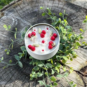 May include: A white candle in a round tin, decorated with red cherry and white flower accents. The candle is surrounded by green foliage, creating a fresh, natural aesthetic. The candle is placed on a wooden surface.