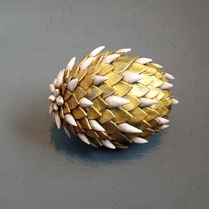 May include: A golden, egg-shaped decorative object with a textured surface. The object is covered in overlapping, scale-like shapes, with white, pointed accents. The item is likely a unique home decor piece.