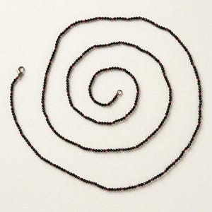 May include: A long beaded necklace arranged in a spiral pattern on a plain surface. The necklace consists of small, faceted black beads interspersed with tiny, vibrant red beads at regular intervals, finished with a silver-toned metal clasp.