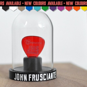 May include: A red guitar pick with the text Gravity JHS Pedals inside a clear glass dome display case. The black circular base features the white text JOHN FRUSCIANTE. The top banner reads NEW COLOURS AVAILABLE with a row of coloured icons.