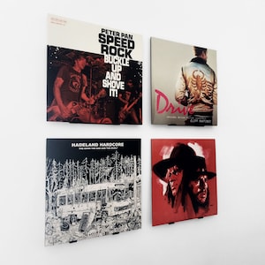 Może przedstawiać: Cztery oprawione okładki albumów wystawione na białej ścianie. Okładki przedstawiają grafiki albumów dla "Peter Pan Speed Rock", "Drive", "Hadeland Hardcore" i czerwoną okładkę z dwiema twarzami. Grafika albumu zawiera tekst i ilustracje.