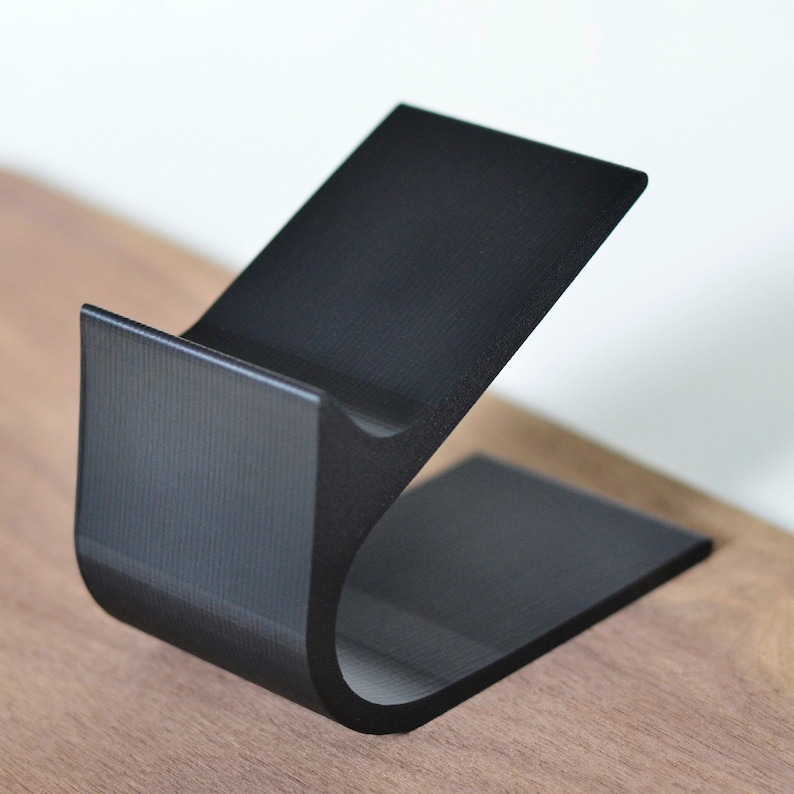 Peut inclure: Un support en plastique noir mat imprim&eacute; en 3D avec un profil incurv&eacute; en forme de C. Le support pr&eacute;sente une base plate, un bras de support vertical et un berceau inclin&eacute; con&ccedil;u pour maintenir un objet, avec une finition de surface textur&eacute;e.