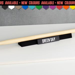 Könnte beinhalten: Ein heller Holztrommelstock auf einem schwarzen, horizontalen Präsentationsständer. Der Ständer trägt den Schriftzug GREEN DAY in weißen Großbuchstaben. Darüber befindet sich ein Banner mit dem Text NEW COLOURS AVAILABLE und farbigen Symbolen.