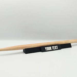 Puede incluir: Una baqueta de madera de color claro apoyada horizontalmente sobre un soporte de exhibición rectangular negro. La parte frontal del soporte presenta el texto blanco YOUR TEXT impreso en una fuente sans-serif, creando un contraste con el negro.