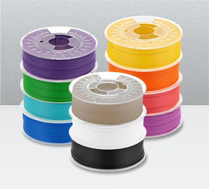 Peut inclure: Une collection de onze bobines circulaires en plastique de filament pour imprimante 3D empil&eacute;es en trois colonnes. Les bobines pr&eacute;sentent diverses couleurs unies, notamment violet, vert, bleu clair, bleu fonc&eacute;, jaune, orange, rouge, magenta, beige, blanc et noir.