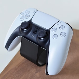 May include: A white and black wireless video game controller resting on a black curved stand. The controller features two black analog sticks, a central touchpad, a PlayStation logo, and four action buttons with geometric symbols on the right side.