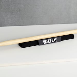 Op de afbeelding: Een zwarte standaard met een zwart-wit bord met de tekst "GREEN DAY" en een houten drumstick erop.