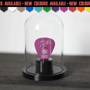 Può includere: Un plettro per chitarra viola con una firma bianca e il logo Planet Waves, esposto all'interno di una cupola di vetro trasparente su una base circolare nera. Sopra, un banner recita NEW COLOURS AVAILABLE con dieci icone pentagonali.