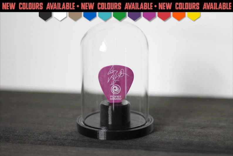 May include: A purple guitar pick with a white signature and the Planet Waves logo, displayed inside a clear glass dome on a black circular base. Above the dome, a banner reads NEW COLOURS AVAILABLE with a row of ten coloured pentagon icons.