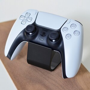 Peut inclure: Une manette de jeu sans fil blanche et noire dot&eacute;e d'un pav&eacute; tactile central, de deux joysticks noirs, d'une croix directionnelle et de quatre boutons d'action avec des symboles g&eacute;om&eacute;triques, pos&eacute;e sur un support noir incurv&eacute; de rangement.